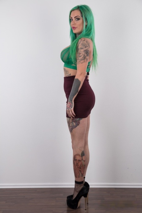 Татуированная девушка с зелеными волосами и проколотыми сосками стоит обнаженной после раздевания