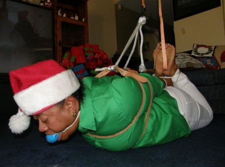 An overweight black woman is tying herself up in her Santa headwear while being ballgged.