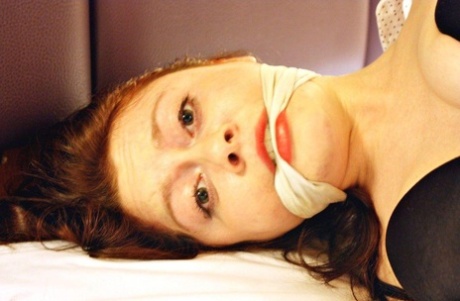 While being hogtied in her clothes on the bed, the natural redhead is left with cramped and gagged.