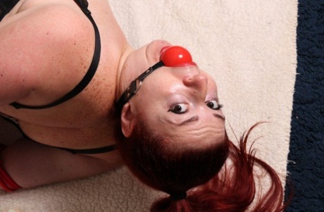 Thick redhead is hogtied in her lingerie while sporting a big ball gag