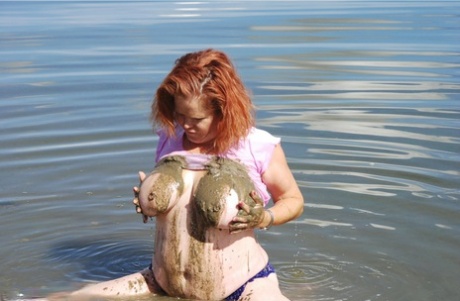While in shallow water, Misha, a redheaded amateur, dips into mud to cover her large tits.