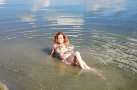 Misha, a redheaded amateur swimmer, dips into mud to cover her large tits in shallow water.