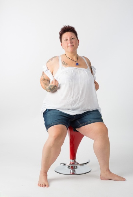 After getting completely naked, a tattooed amateur gets up and sits on top of a stool.