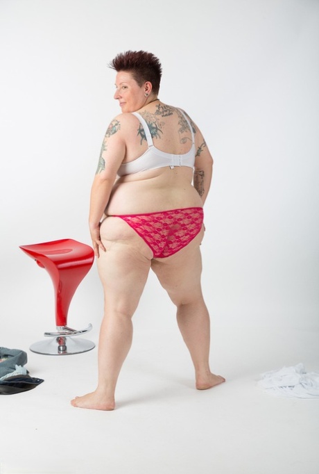 A tattoo girl who is an older woman gets naked and sits on a stool, looking rather uncomfortable.