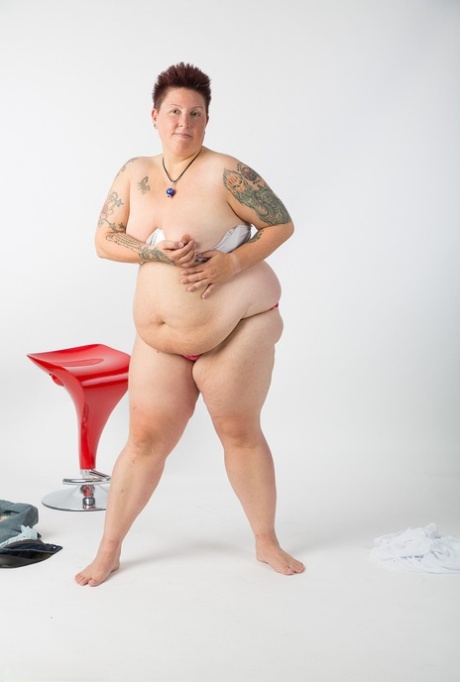 The naked tattoo girl, who is an older woman than the average, sits on a stool.