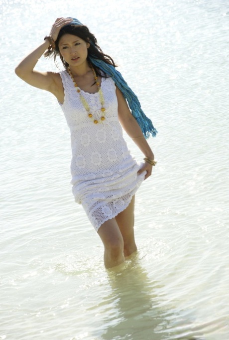 Donning a white dress, an Asian girl walks into the ocean with her knees raised.