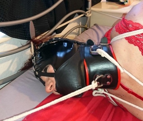 A white girl with a collar is tied down to a bed in red lingerie and a latex hood.