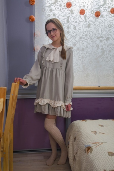 On her bed, the nerdy girl removes her dress and accessories while wearing glasses.