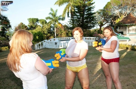 Women with large tattoos soak white t-shirts in the yard with water guns that are super-soaking.