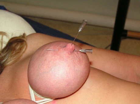 A woman who is blonde has her breasts and nipples pinched with needles.