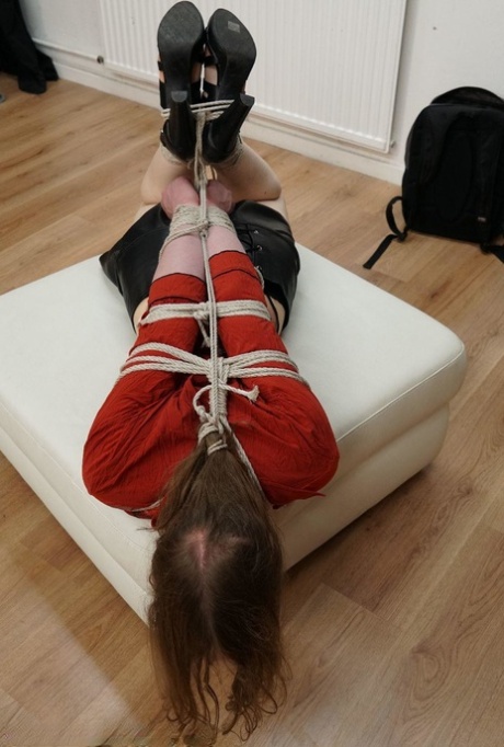 A white woman is fully clothed while hogtying herself on an ottoman.