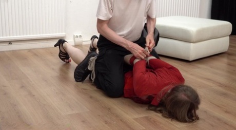 In a fully clothed position, the Caucasian female is hogtied over an ottoman.
