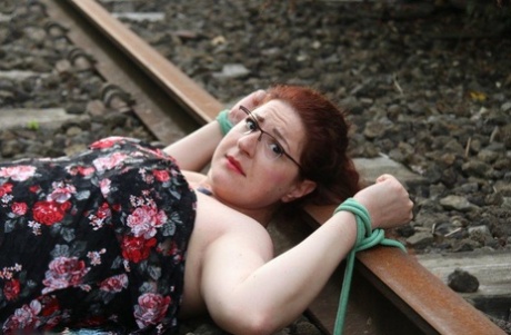 Wearing a dress and glasses, the natural redhead is tied to train tracks.