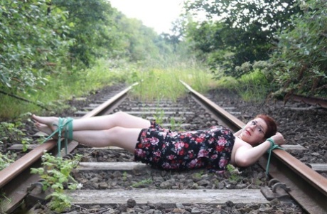 The natural redhead is wearing a dress and glasses and has been tied down to the train tracks.