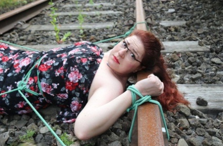 Wearing a dress and glasses, the natural redhead is tied to train tracks.