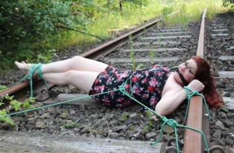 The natural redhead is wearing a dress and glasses and has been tied down to the train tracks.