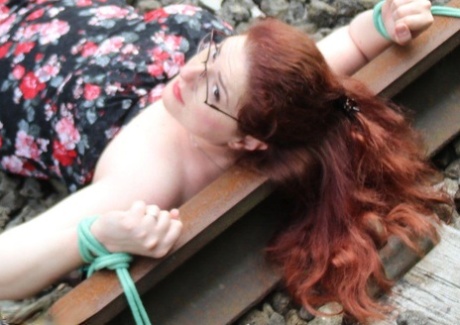 Wearing a dress and glasses, the natural redhead is tied to train tracks.