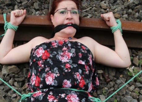 The natural redhead is wearing a dress and glasses and has been tied down to the train tracks.