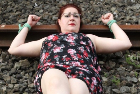 Wearing a dress and glasses, the natural redhead is tied to train tracks.