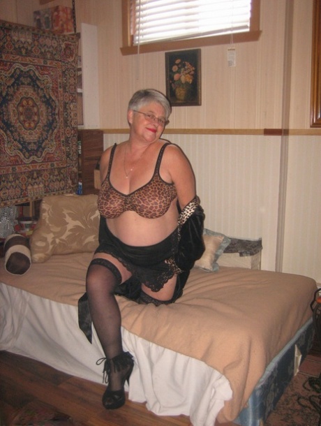 On her bed, the granny Girdle Goddess with silver hair and stockings engages in masturbation.