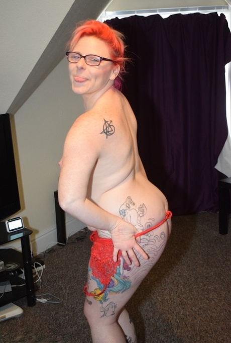 A tattooed photographer removes their red lingerie to capture a nude photo while wearing glasses and heels.
