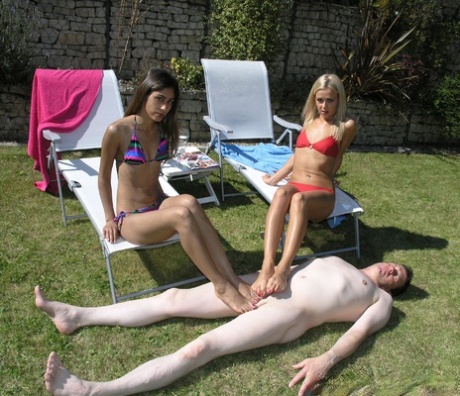 In a backyard, girls wearing bikinis place their bare feet on a naked man's penis.