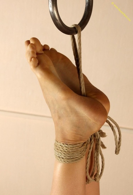 Using ropes, the naked female is restrained at her unclothed feet by wrapping them around her ankles.
