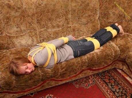 Barefoot woman struggles on couch, her clothes tied tightly as she tries to get back on.