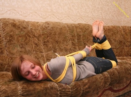 As the barefoot female on the couch struggles to move, she is closely tied up with her clothes hanging over her back.