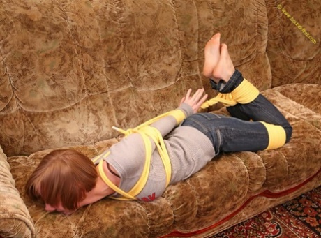 As the barefoot female on the couch struggles to move, she is closely tied up with her clothes hanging over her back.