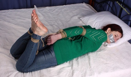 A white girl who is barefoot is silenced by duct tape while she is tied up in her clothes.