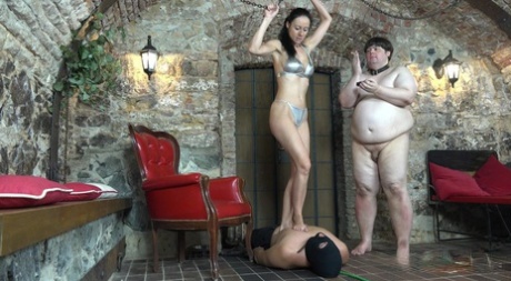 The cruel woman stomps on the back of an armoured male slave while using a fat man as an ashtray.