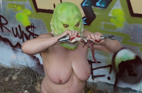 While wearing a costume mask, Speedy Bee, a naked British woman, consumes a fish.