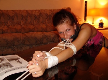 White girl sporting cleave gag while restrained with rope draped over coffee table.