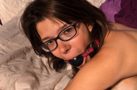 A gagged girl is pinned down to the bed with all fours while wearing glasses and a thong.