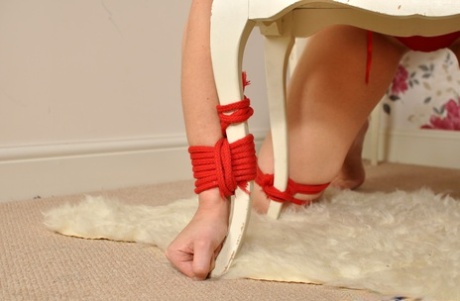 The nude blonde performs a ball gag while being held to the ground on a footstool.