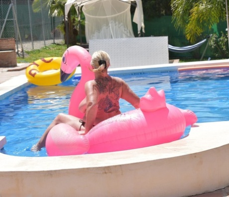 An inflatable in a pool is used by Melody, an older tattooed blonde, who is naked on top.