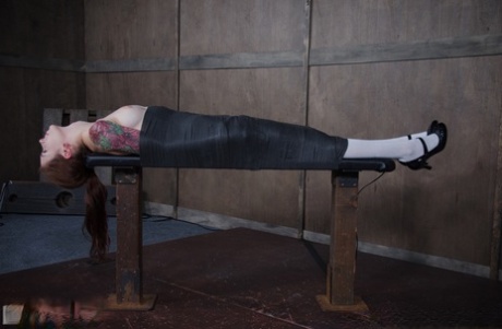 An individual with tattoos, Anna De Ville, is subjected to a throat fuck on the bondage table.