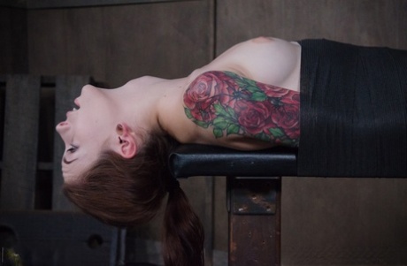 The throat fuck on a bondage table is being subdued by Anna De Ville, a woman with tattoos.