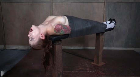 The throat fuck on a bondage table is being subdued by Anna De Ville, a woman with tattoos.