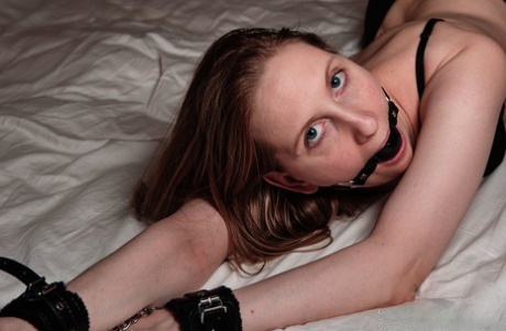 A black bra and panties are worn by a natural redhead who is ball gagged and cuffed on her bed.