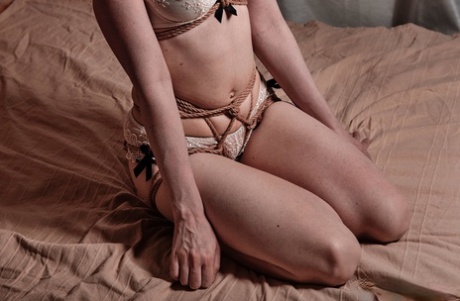 A white woman wearing a bra and panty ensemble on her bed, tied up with a rope.