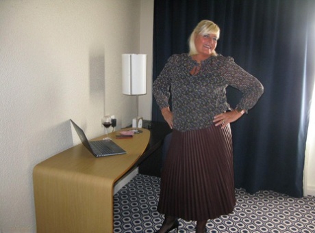 At a desk, an adult BBW Chrissy Uk displays her breasts and legs while sipping wine.