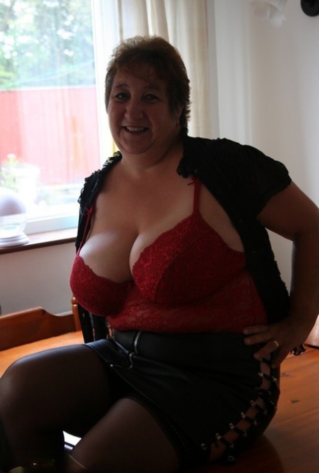 An obese and older woman named Kinky Carol is holding her massive breasts near a table.