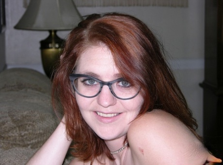 The young redhead with nipple piercings exposes herself while wearing glasses.