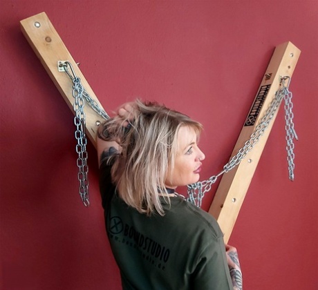 A St Andrew's Cross is the setting for Roxxxi Manson, a blonde with hair who removes a ball gag.