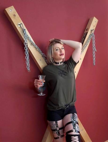 The ball gag is disregarded by Roxxxi Manson, who is blonde and stands up in front of a St Andrew's Cross.