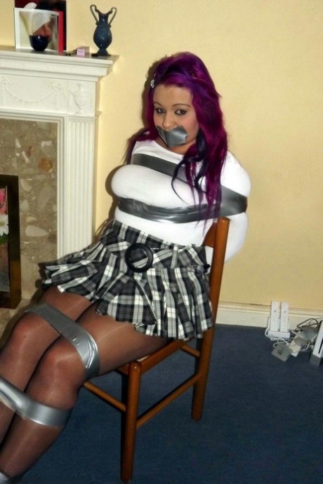 Busy girl, pink in hair, is gagged while restrained to a chair.