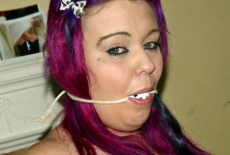 An energetic female with a strong chest and pink hair experiences being held down in an armchair while being gagged.
