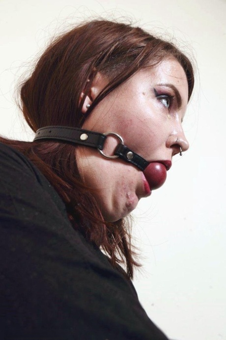 While wearing a ball gag and being bound with straps, Luna La Roux, the tattooed redhead, is shown.